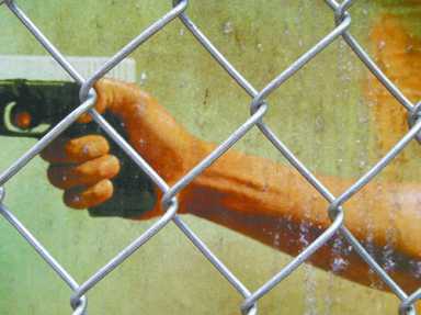 A hand holding a handgun, viewed through a chain-link fence.