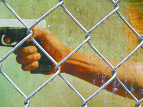 A hand holding a handgun, viewed through a chain-link fence.
