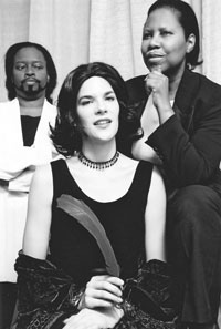 A person in a dark dress and necklace sits in the foreground, with two other people behind them.
