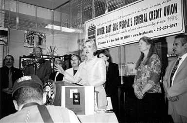 Black and white image of a woman speaking at a podium with a banner behind her, surrounded by other people.