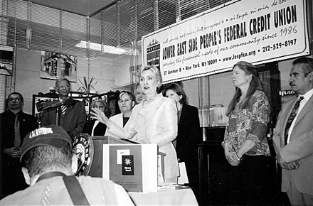 Black and white image of a woman speaking at a podium with a banner behind her, surrounded by other people.