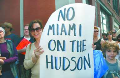 A woman in sunglasses holds a protest sign that reads "NO MIAMI ON THE HUDSON."