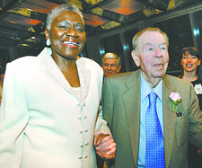 An older woman in a light suit and an older man in a brown suit and blue tie smile and hold hands.
