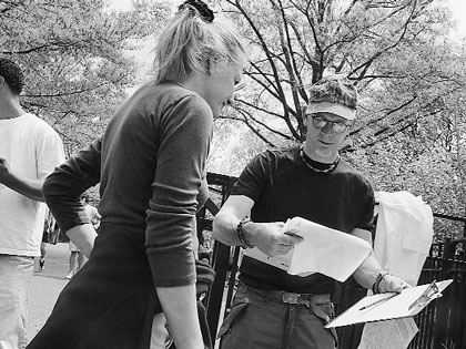 Black and white image of two people, one appearing to be a woman with hair tied up, and a man in a cap and glasses holding papers.