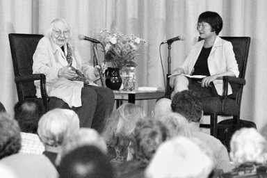 Black and white image of two women, one older and one younger, seated on a stage with microphones, facing an audience.