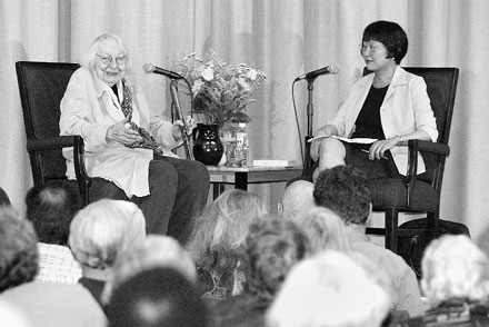 Black and white image of two women, one older and one younger, seated on a stage with microphones, facing an audience.