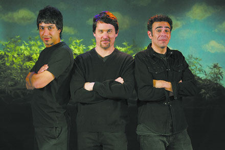 Three men in dark clothing stand with crossed arms against a painted backdrop.