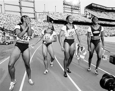 Black and white image of four women in "USA" track uniforms celebrating on a track in a stadium.