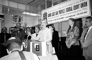 Black and white image of a woman speaking at a podium with a banner behind her, surrounded by other people.