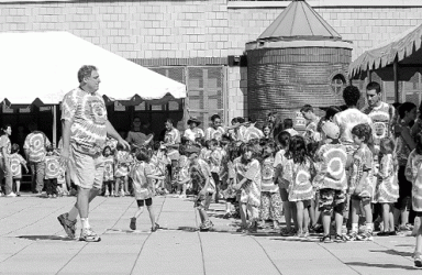 A group of children in matching patterned outfits line up outdoors, with an adult supervising.