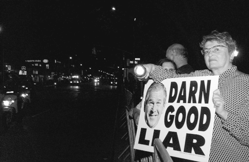 A woman holding a protest sign featuring George W. Bush and the words "DARN GOOD LIAR" at night.