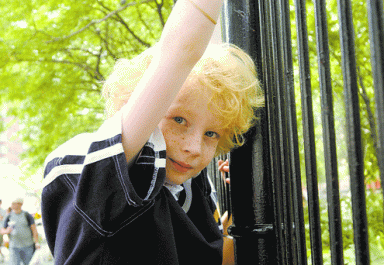 A young child with curly blonde hair and freckles peeks from behind a dark fence.