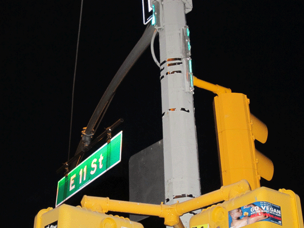 Photo by Lincoln Anderson At some point, a street co-naming sign for “Jodie Lane Place,” as well as signs for E. 11th St. and First Ave., were hacksawed off this lamppost, while a large, new, cantilever sign for First Ave. was added. Some jagged thin green strips, remnants of the former signs, are still visible toward the top of the photo.