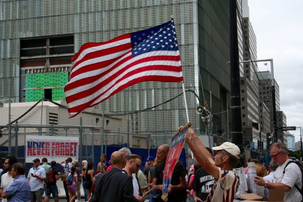 In front of the PATH station on Vesey St., a man was waving an American flag and holding a sign reading "In God We Trust" on Friday. Downtown Express Photo by Yannic Rack.