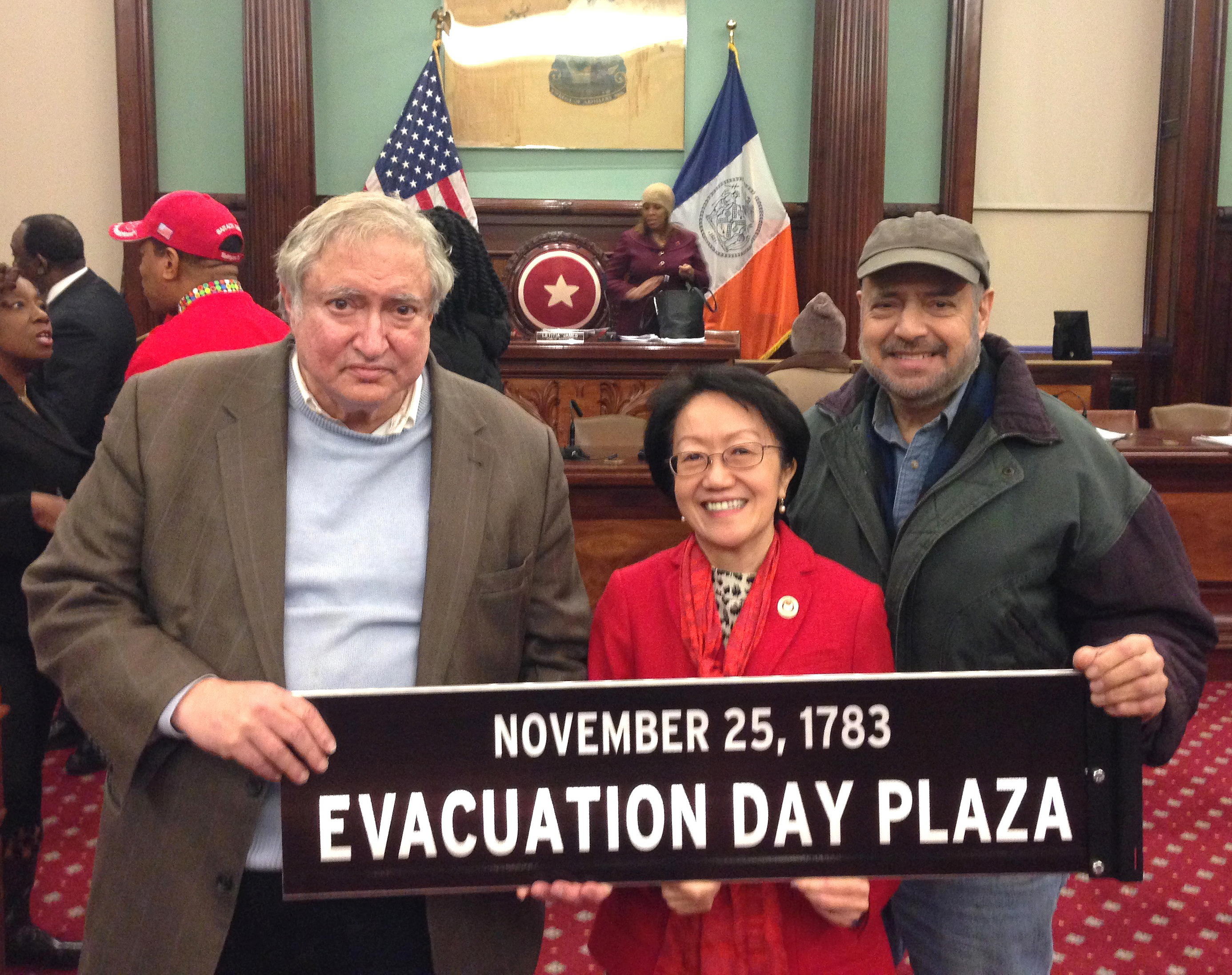 Photo courtesy of Arthur Piccolo Downtown’s Councilmember Margaret Chin, center, beams in the Council chamber as she holds up the “Evacuation Day Plaza” sign just approved for Bowling Green, flanked by Lower Manhattan Historical Society co-founder James Kaplan on the left, and Bowling Green Association head Arthur Piccolo on the right.