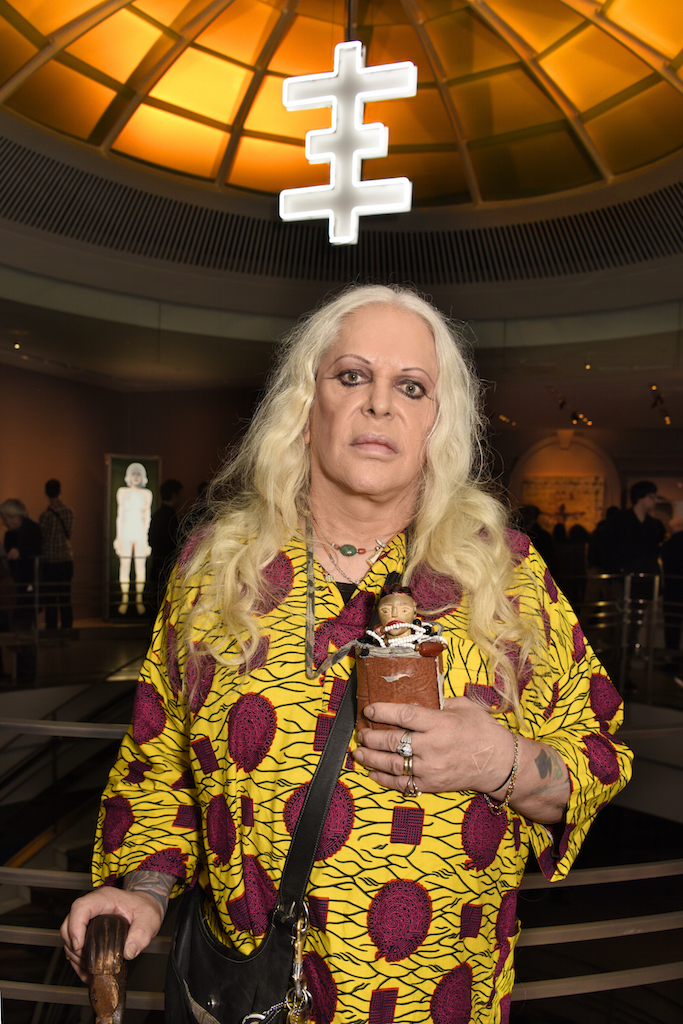 “Pandrogenic” artist Genesis Breyer P-Orridge at the opening of her/his show at the Rubin Museum in Chelsea. Above is P-Orridge’s “psychic cross.” Glowing in the background is an image that blends P-Orridge and her/his late partner Lady Jaye. P-Orridge is holding a voodoo doll representing Lady Jaye.  Photo by Bob Krasner