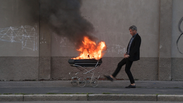 Maurizio Cattelan kicks a burning baby carriage in Milan, in “Maurizio Cattelan: Be Right Back.” Photo courtesy Maurizio Cattelan Archive.