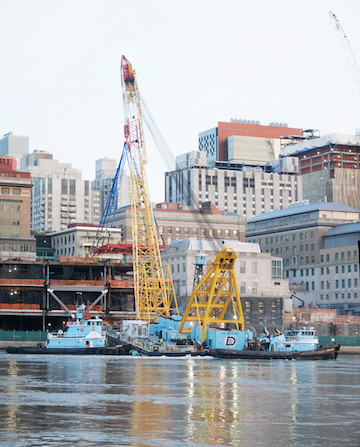 A daytime shot of the crane, with the modules already inserted just ahead of the crane and the FDR Drive at the green fencing level just above the river. | FRANK FARANCE 