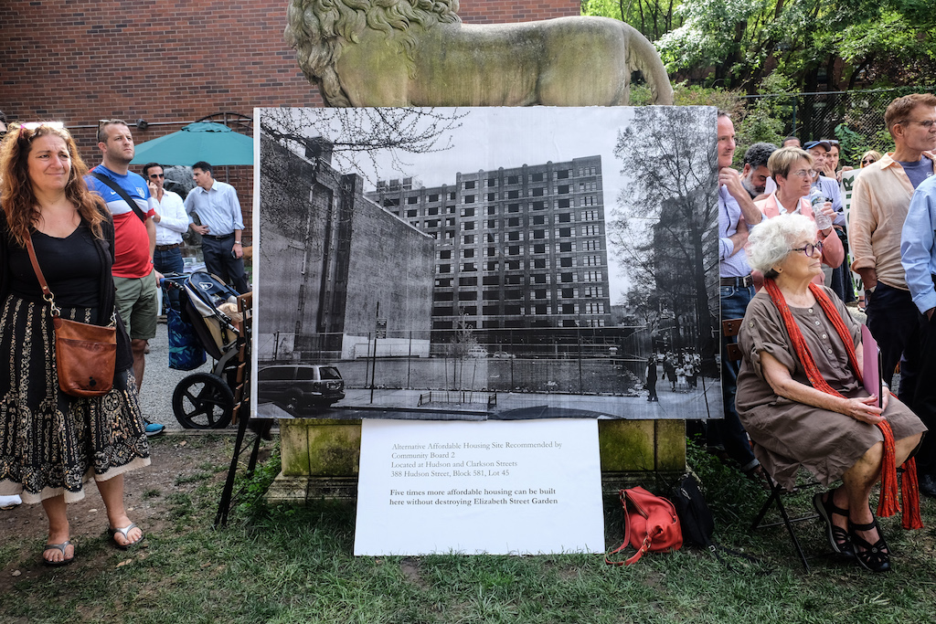 At a rally to save the Elizabeth St. Garden two weeks ago, Sharon D'Lugoff, left, daughter of the late Village Gate impresario Art D'Lugoff, stood next to a photo of the long-vacant city-owned lot on Hudson St. that C.B. 2 is urging the de Blasio administration ho use for a housing project instead of the garden. The blown-up photo was resting against one of the garden's signature lion monuments. However, de Blasio on Tuesday announced a plan to develop the Hudson St. lot as a park, with the possibility of some affordable housing there, as well. File photo by Tequila Minsky