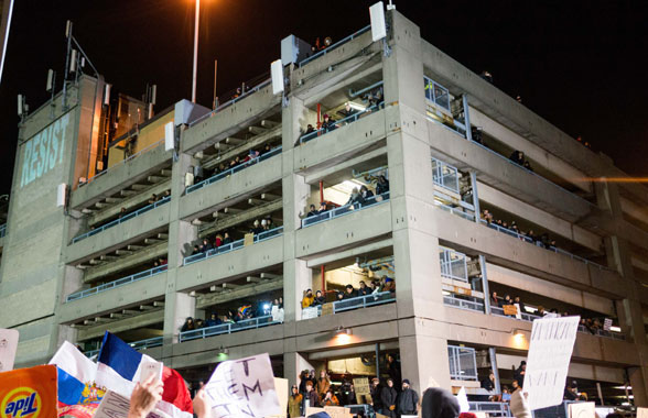 Ban on the Run: Protests at JFK, Jubilation at Brooklyn Courthouse 4 Protestors in a parking lot at the airport on which the word “Resist” was illuminated. Photo by Julius Constantine Motal.