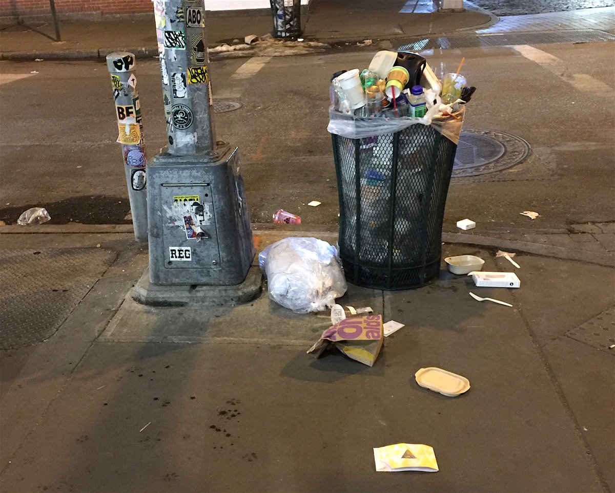 An all-too-familiar scene in Soho nowadays: a street corner with an overflowing garbage can surrounded by litter on the sidewalk.