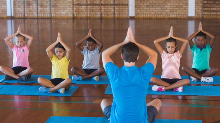 School kids and teacher meditating during yoga class in a school gym.