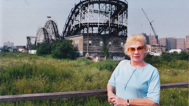 Mae Pompano standing on the boardwalk in Coney Island. In the background is the house that she lived in for 40 years, which is under the Thunderbolt roller coaster.