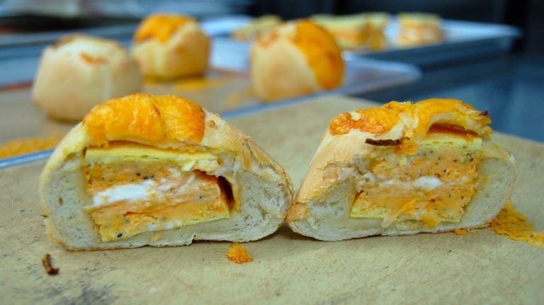 Milk Bar's egg & cheddar menu item has been re-imagined. Say goodbye to the croissant-like bread and hello to the Bomb. Behold: the Egg & Cheddar Bomb, sliced in half to display the egg, cheddar cheese and Sriracha cream cheese. It's topped with more cheese and julienned Spanish onions. 