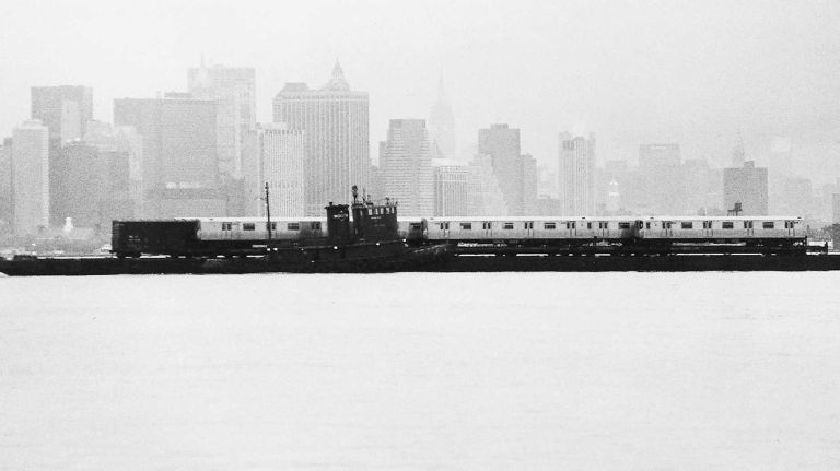 A barge tows refurbished subway cars across lower New York Harbor toward Brooklyn at dusk. According to the Transit Authority Public Affairs Office, the subway cars are refurbished in Hornell, New York, then shipped back from the western part of the state to Manhattan, the last leg by barge across the Harbor. These cars were newly refurbished and ready to be put into service. (May 1, 1991)