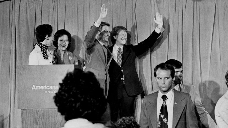 Walter Mondale and Jimmy Carter wave to the crowd at the Americana Hotel on July 16, 1975, after receiving their nomination at the Democratic National Convention. Standing behind the podium are their respective wives, Joan Mondale and Rosalynn Carter.