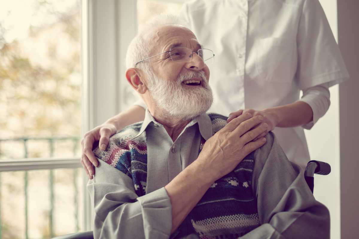 Senior man sitting on the wheelchair, laughing and holding his nurse’s hand