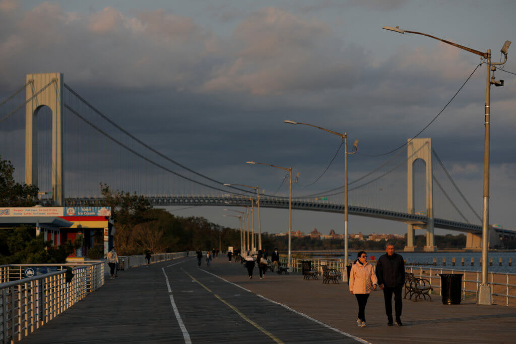 People walk along the Franklin D Roosevelt Boardwalk in front of the Verrazzano-Narrows Bridge on Staten Island in New York City