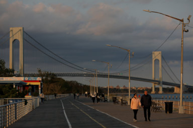 People walk along the Franklin D Roosevelt Boardwalk in front of the Verrazzano-Narrows Bridge on Staten Island in New York City