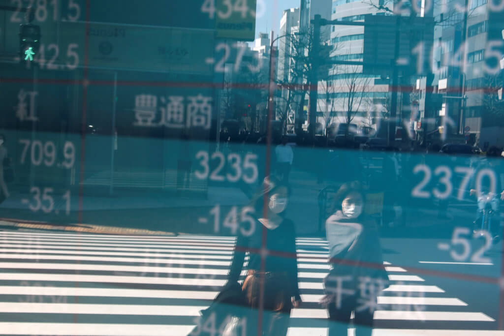 People wearing protective face masks, following an outbreak of the coronavirus, are reflected on a screen showing Nikkei index, outside a brokerage in Tokyo