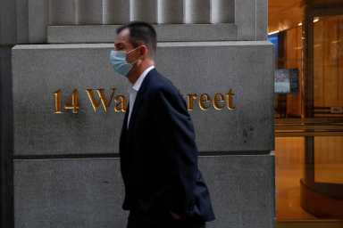 A man wearing a protective face mask walks by 14 Wall Street, as the global outbreak of the coronavirus disease (COVID-19) continues, in the financial district of New York