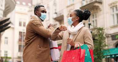 Portrait of cheerful African American young man and woman in medical masks with xmas presents meeting on street after holiday shopping and kindly speaking while snowing. Friends greeting with elbows