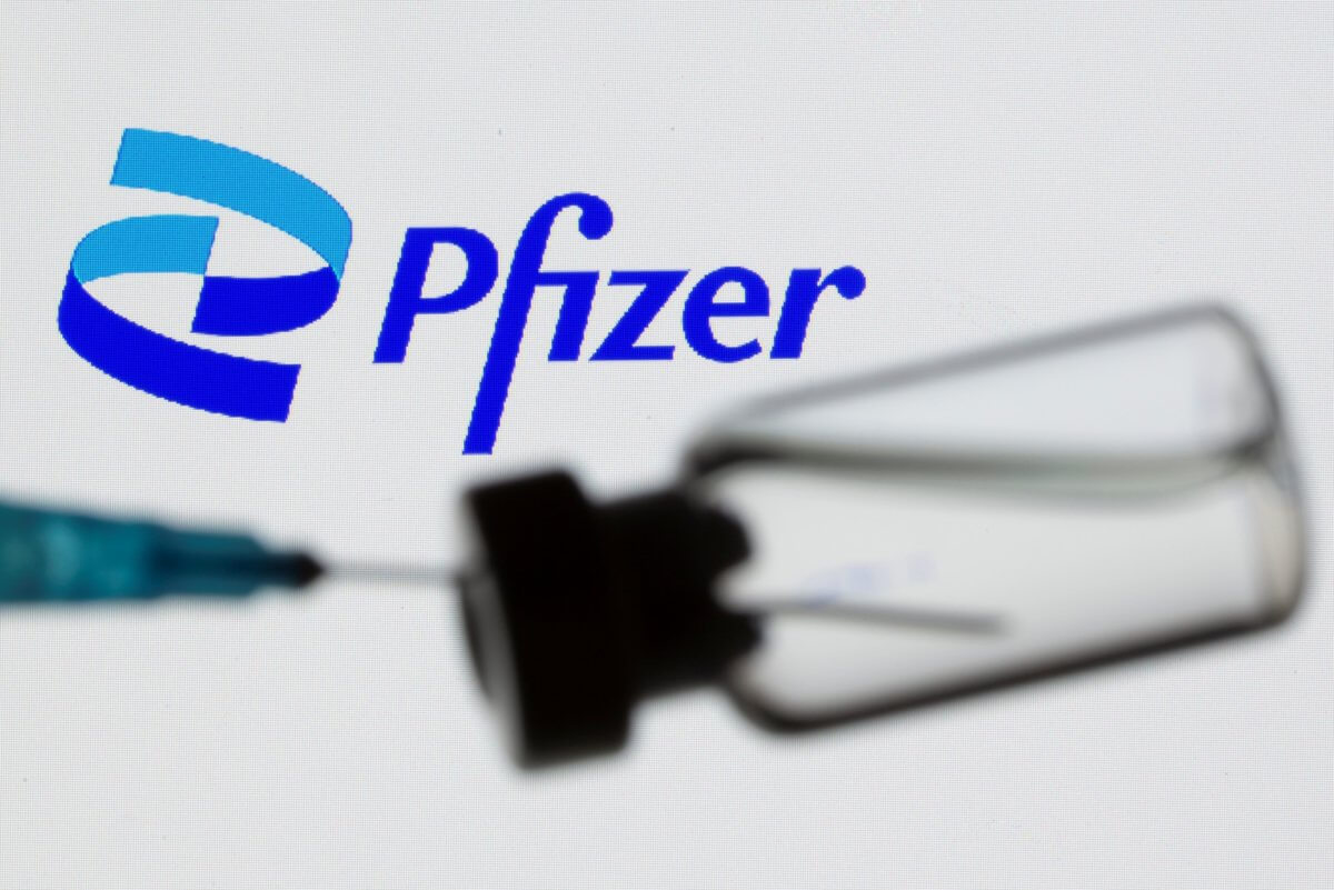 Syringe and vial are seen in front of displayed new Pfizer logo in this illustration taken