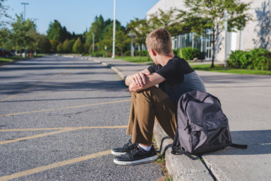 Lonely teenage boy sitting on curb next to high school.