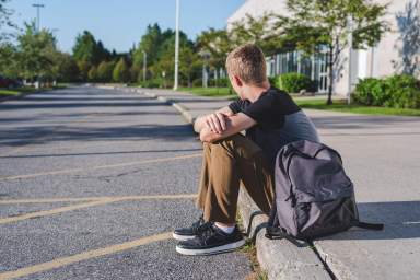Lonely teenage boy sitting on curb next to high school.