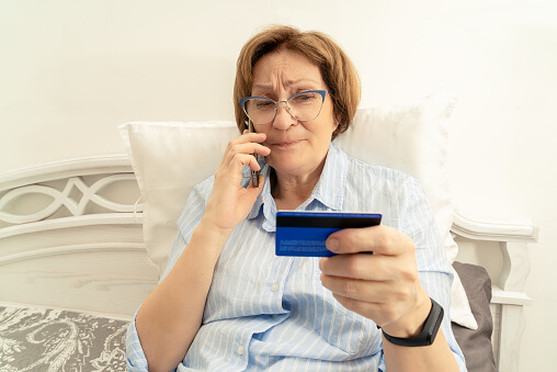 Elderly doubtful woman talking on a mobile phone and holding a bank card in her hand
