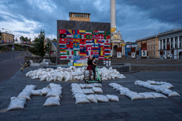 Women ride a scooter through Kyiv's Maidan Square, past sandbags that spell out 'HELP,' and flags displayed from around the world, in Ukraine after the Russia invasion.