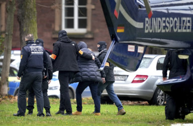 An suspect, second right, is escorted from a police helicopter by police officers after the arrival in Karlsruhe, Germany, Wednesday, Dec. 7, 2022. Thousands of police officers carried out raids across much of Germany on Wednesday