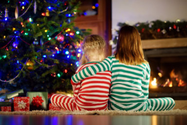Happy little sisters wearing Christmas pajamas opening gift boxes by a fireplace in a cozy dark living room on Christmas eve.