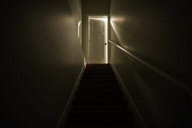 A dark stairwell with illuminated by a slightly opened door at the top of the stairs.