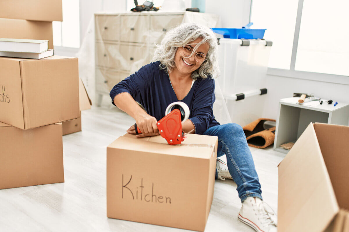 Middle age grey-haired woman smiling happy packing kitchen cardboard box at new home.