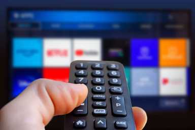 A man is holding a remote control of a smart TV in his hand. In the background you can see the television screen with streaming entertainment apps for video on demand