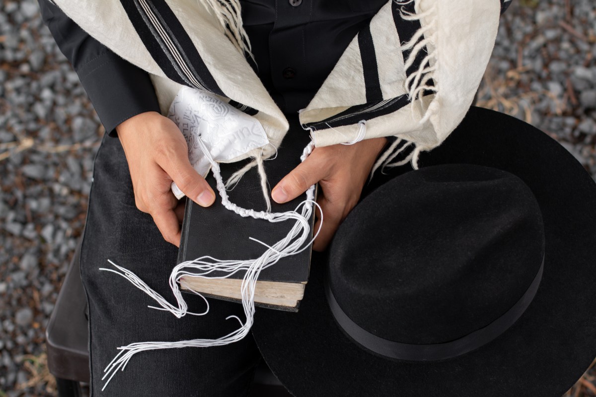 Close-up of a Jew holding a siddur on which rests the tzitzit of his tallit and next to it a hat.