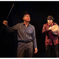 two people, one holding a drum, performing on stage in an off-Broadway show