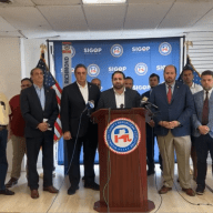 group of men and women inside a building at a podium speaking about attempted assassination of Donald Trump
