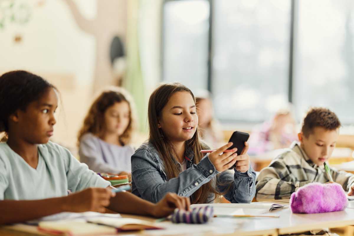 Smiling elementary student using mobile phone while having a class with her friends at elementary school.